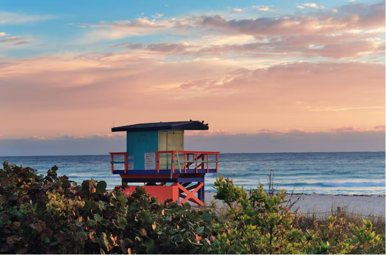 A colorful lifeguard tower stands on a beach with the ocean in the background under a pastel sunset sky.