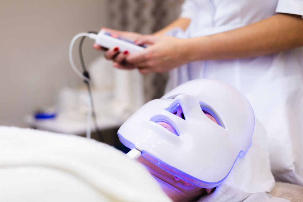 A client wears an LED light therapy mask while a beautician in a white uniform holds the control device.