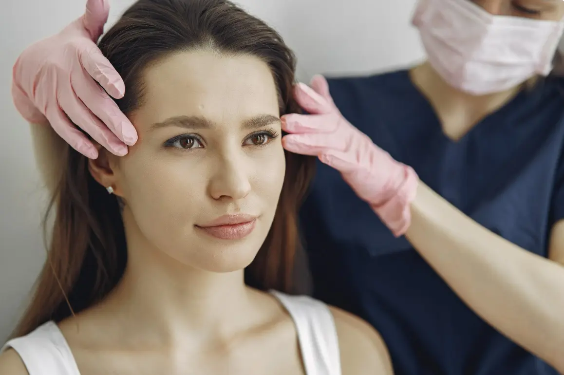 A skincare professional wearing gloves gently examines a woman's face during a facial aesthetics consultation in a modern clinic.